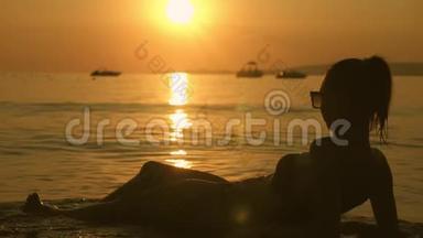 日落时分，美丽的年轻女孩躺在海滩上。 度假的女孩在海滩上放松，看日落。 夏季